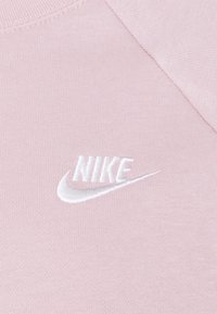 Ljusrosa tyg med en vit broderad Nike-logotyp på bröstet. Mjuk textur med rund halsringning och raglanärm.