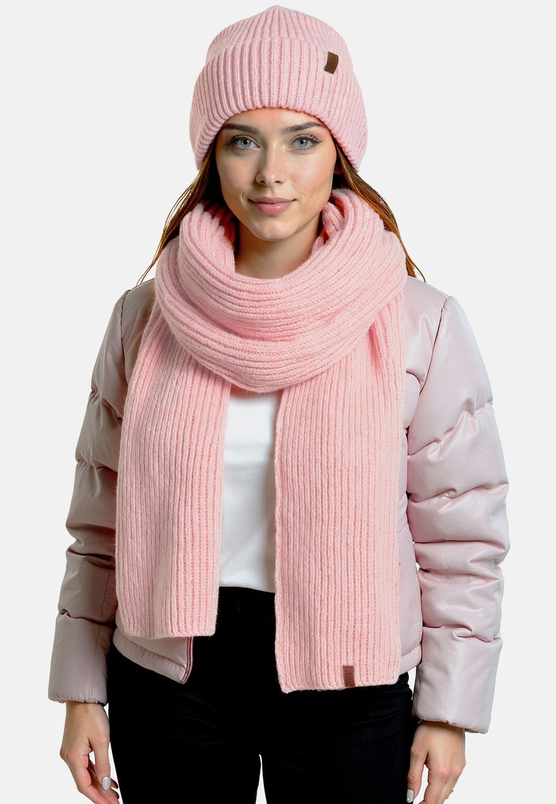 Gorro de punto rosa y bufanda a juego con textura acanalada, llevados con una chaqueta acolchada rosa claro. La bufanda está enrollada alrededor del cuello.