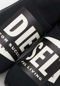 Mustat kangas liukupohjaiset kengät, joissa on rohkeat valkoiset kirjaimet "DIESEL" ja "FOR SUCCESS" sivuilla, ja sileä, teksturoitu pinta.