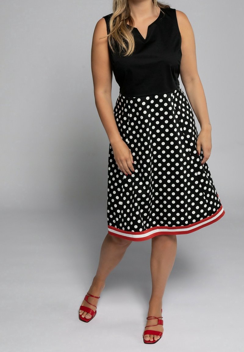 Femme portant un top noir sans manches et une jupe noire mi-longue à pois blancs avec un ourlet rayé rouge, associée à des sandales rouges ouvertes aux orteils.