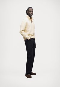 Pull crème à fermeture éclair, pantalon noir, chaussures marron. Le pull présente un col haut et une texture subtile, avec un design minimaliste.