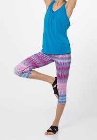 Person som bär en blå linne och mönstrade leggings, balanserar på ett ben med armarna uppsträckta, och visar en yoga pose.