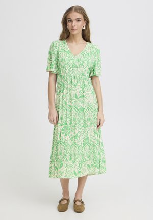 Tânără purtând o rochie midi verde cu alb, cu mâneci scurte, și pantofi maro fără toc, stând în picioare pe un fundal gri simplu.