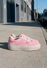 Ζευγάρι ροζ sneakers με πλατφόρμα και λευκά κορδόνια τοποθετημένα σε ένα ηλιόλουστο αστικό πεζοδρόμιο κοντά σε παρκαρισμένα αυτοκίνητα και κτίρια.