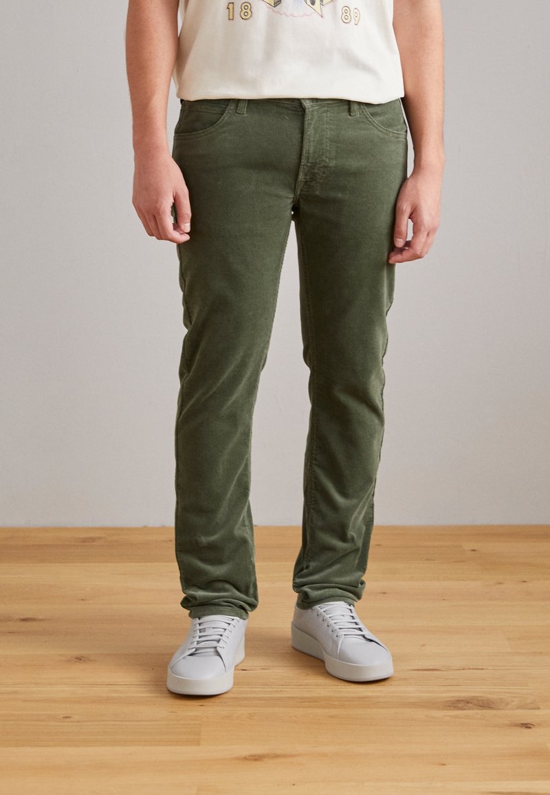 Pantalones de pana verde oliva con un ajuste slim, que cuentan con una cinturilla estándar y cinco bolsillos, combinados con zapatillas blancas.