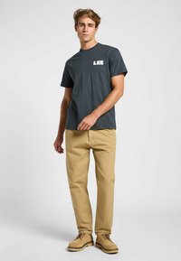 Grå bomullströja med "LEE"-logotyp i vitt, kombinerad med ljusbeige chinos och bruna stövlar. Avslappnad passform och korta ärmar.