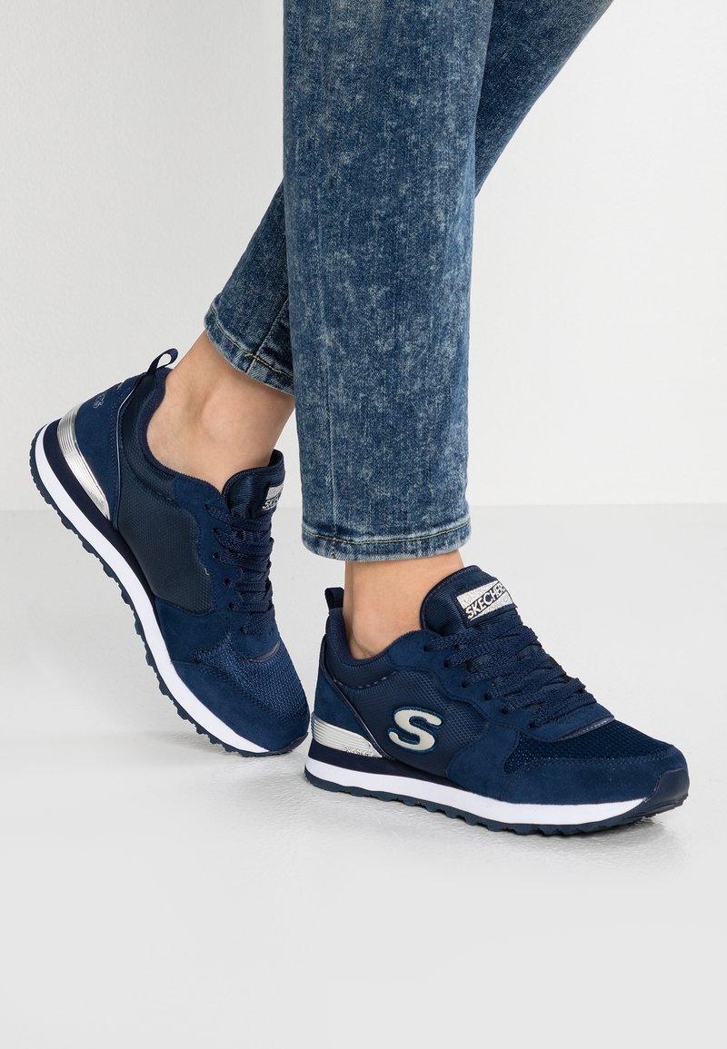 Person som bär mörkblå Skechers-skor och blå denimjeans, står på en ljusgrå golv mot vit bakgrund.