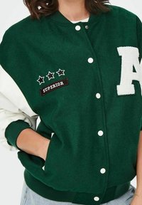 Zielona i biała kurtka w stylu varsity o miękkiej teksturze, z białą literą "A", trzema gwiazdkami oraz naszywką "SUPERIOR". Zapięcie na zatrzaski z przodu.