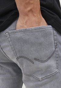 Vaqueros de mezclilla grises con un bolsillo trasero que presenta detalles de costura curva. El material es suave con una ligera textura. Combinados con una camisa negra.