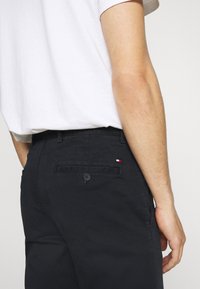 Black cotton trousers with a back pocket featuring a small red, white, and blue logo. Pants have a straight fit and a button closure.