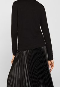 Černý pletený svetr s dlouhými rukávy, zdobený žebernou strukturou a knoflíky na zápěstí. Kombinován s černou plisovanou sukní.
