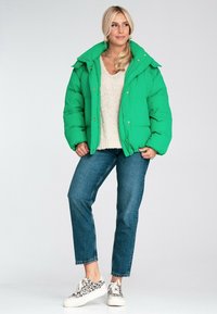 Jasnozielona kurtka puchowa z wysokim kołnierzem i złotymi detalami, noszona na kremowym swetrze, w zestawie z dżinsami i trampkami w panterkę.