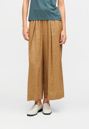 Pantalon marron clair à jambes larges en tissu texturé avec des plis à l'avant, associé à des chaussures blanches à enfiler et un haut turquoise.