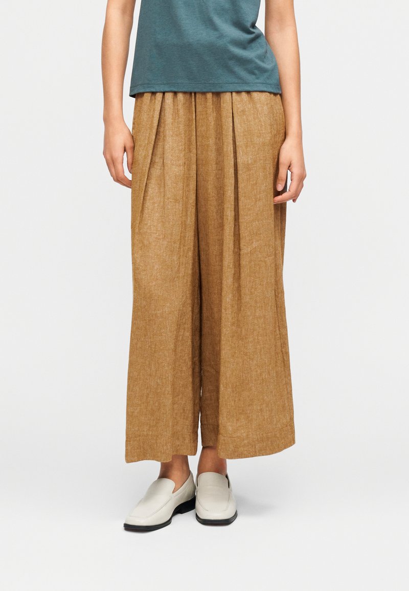 Pantalon marron clair à jambes larges en tissu texturé avec des plis à l'avant, associé à des chaussures blanches à enfiler et un haut turquoise.