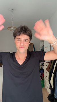 Mladý človek oblečený v čiernom tričku s výstrihom do V, zdvihol obidve ruky. Krátke, kučeravé vlasy a mierny úsmev, s pekne usporiadaným pozadím.