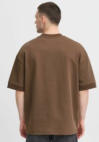 Brun oversized t-skjorte med korte ermer, rund hals og glatt tekstur. Bakvinduet viser ingen mønstre eller logoer, enkel design.