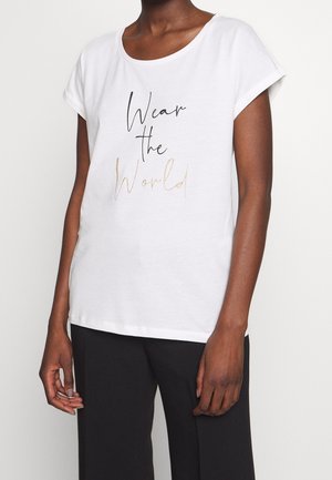 Osoba nosząca białą koszulkę z napisem "Wear the World" w czerni i złocie, połączoną z czarnymi spodniami, na jednolitym tle.