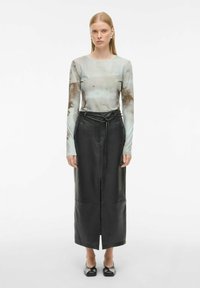 Naine kannab läbipaistvat pika varrukaga pluusi pruuni mustriga, musta kõrge vöökohaga nahkse seeliku koos vöödiga ning musti ruudukujuliste varvastega kõrgemaid kingi.