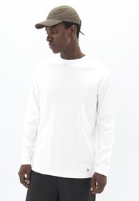 Vyras, dėvintis smėlio spalvos beisbolo kepuraitę, baltą ilgomis rankovėmis marškinį su mažo logotipo prie apykaklės ir juodas kelnes, žiūri žemyn į kairę.