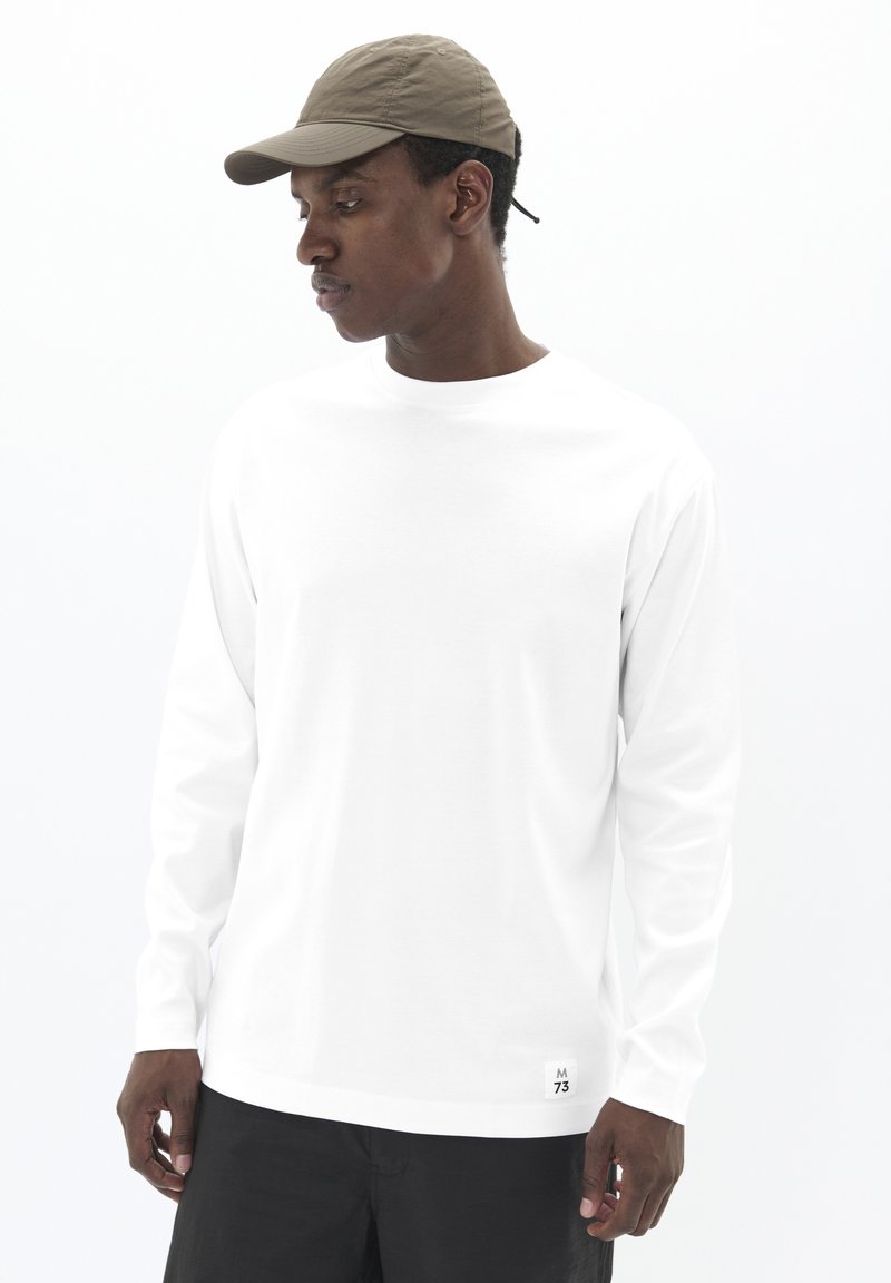 Vyras, dėvintis smėlio spalvos beisbolo kepuraitę, baltą ilgomis rankovėmis marškinį su mažo logotipo prie apykaklės ir juodas kelnes, žiūri žemyn į kairę.