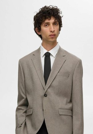 Lys brun, strukturert blazer med slagkrage, enkel knappeåpning og to frontlommer, kombinert med en hvit skjorte og prikkete slips.