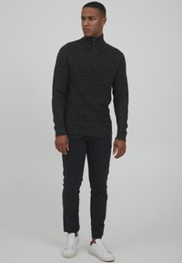Dunkelgrauer Strickpullover mit hohem Kragen und halbem Reißverschluss, kombiniert mit schmal geschnittenen dunkelblauen Hosen und weißen Sneakers, die ein strukturiertes Muster zeigen.
