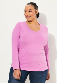 Růžová dlouhá tričko s výstřihem do V, vyrobené z hladké tkaniny, spárované s tmavými džíny. Tričko má přiléhavý střih a jemnou strukturu.