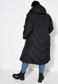 Svart puffjacka med en chevronmönster, huva och hel längd. Kombinerad med blå jeans och kraftiga svarta stövlar.