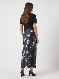 Ženska z dolgimi rjavimi lasmi, oblečena v črno majico s kratkimi rokavi, temno cvetlično midi krilo in črne pete, stoji obrnjenona stran.