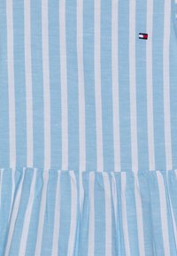 Světlomodré šaty s bílými svislými pruhy, s nabíranou sukní a malým vyšitým logem v horním rohu. Bavlněný materiál.