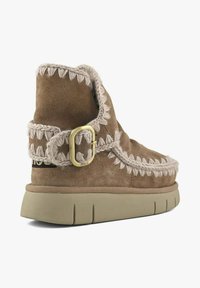 Bottes basses en daim marron avec coutures crème, une boucle décorative et une semelle en caoutchouc texturée. Caractérisées par un bout arrondi et une surface extérieure douce.