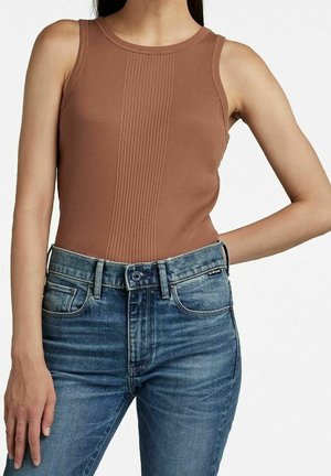 Frau trägt ein ärmelloses braunes, geripptes Top, das in eine blaue, hoch taillierte Jeans mit Knopf- und Reißverschluss gesteckt ist.