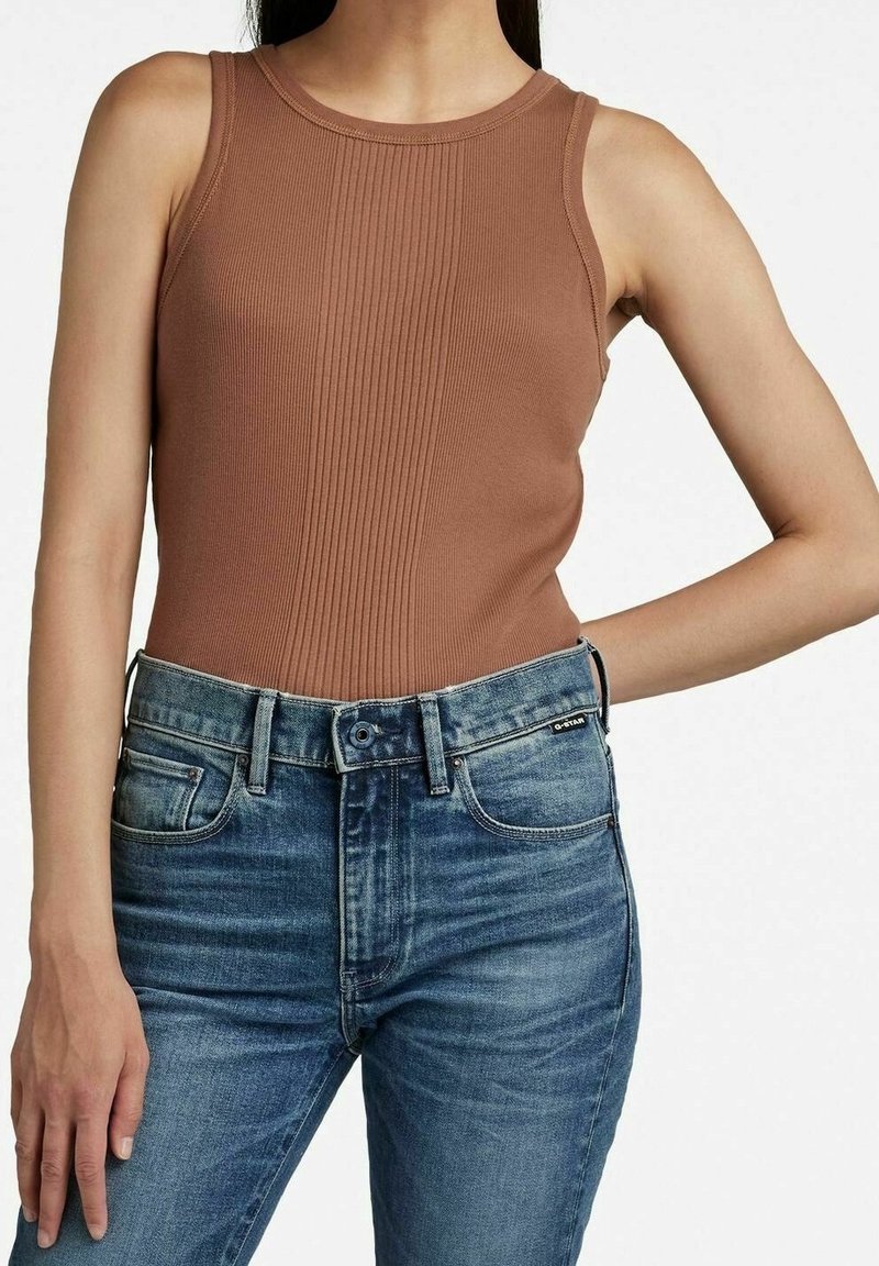 Femme portant un haut sans manches marron côtelé, rentré dans un jean en denim bleu taille haute avec une fermeture à bouton et fermeture éclair.