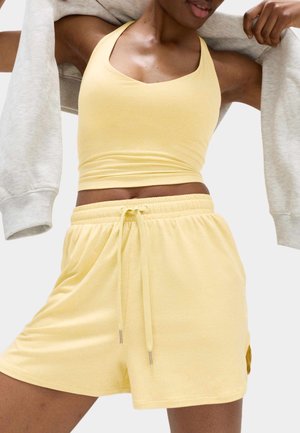 Kvinde iført gul tanktop og matchende snøre-shorts, der holder en grå hættetrøje over skuldrene mod en hvid baggrund.