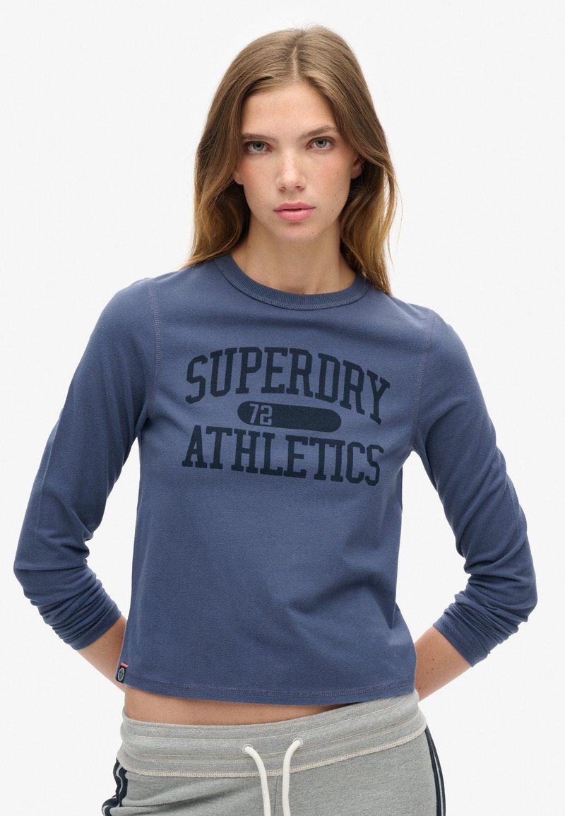 Blauwe sweatshirt met lange mouwen en de tekst "SUPERDRY ATHLETICS" in donkerblauw gedrukt, met een ronde halslijn en een cropped snit. Zachte stof.