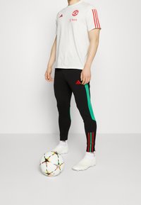 Bílá tričko s krátkým rukávem Adidas s červenými akcenty a logem, v kombinaci s černými legínami s zelenými bočními pruhy a červeným brandingem. Bílý fotbal.