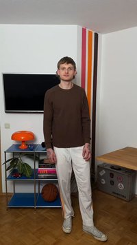 Νέος άνδρας με τατουάζ στέκεται σε ένα δωμάτιο με μια μπάλα του μπάσκετ, πορτοκαλί λάμπα, τηλεόραση, μεταλλικό ράφι και ριγωτή διακόσμηση τοίχου.