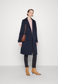 Trench coat bleumarin, cu două rânduri de nasturi și epoleți pe umeri, purtat peste un pulover roz deschis, asociat cu blugi închiși la culoare și cizme scurte bej.