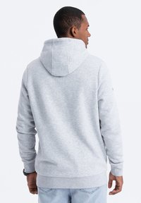 Sudadera gris hecha de tela suave, con capucha, mangas largas y un ribete en el dobladillo. La parte trasera presenta un diseño limpio y sin costuras.