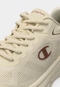 Μπεζ αθλητικό παπούτσι Champion με υφασμάτινη υφή, μπεζ κορδόνια, καφέ λογότυπο "C" στο πλάι και κείμενο "Champion" στη γλώσσα.