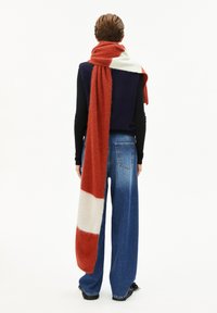 Personne portant une longue écharpe rouge et blanche, un haut sans manches bleu marine sur une chemise à manches longues noire, et un jean large bleu, de dos.