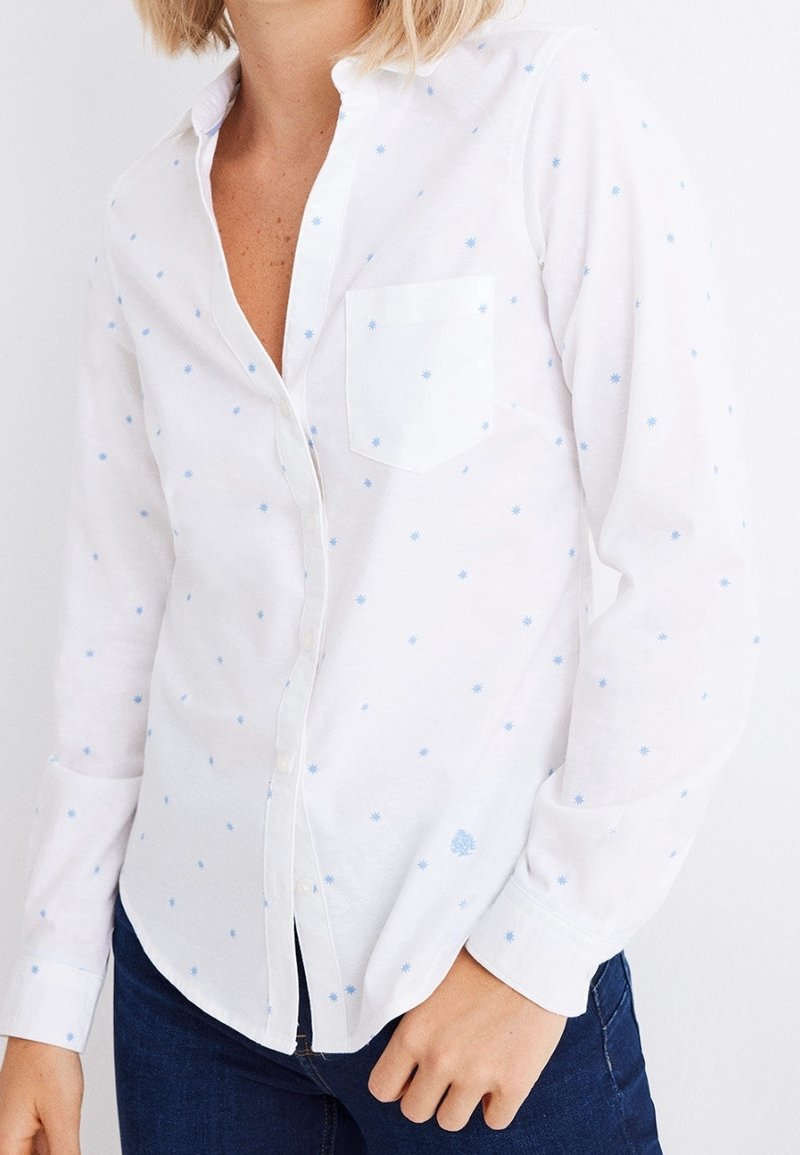 Femme portant une chemise blanche à manches longues boutonnée avec de petits motifs d’étoiles bleues et un jean bleu foncé sur un fond uni.