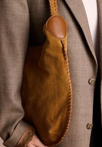 Brown suede shoulder bag with a soft texture, featuring a stitched leather accent and rounded edge, held against a grey textured blazer.