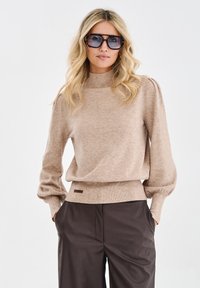 Beżowy sweter dzianinowy z wysokim kołnierzem i balonowymi rękawami, w połączeniu z brązowymi skórzanymi spodniami. Zawiera małą metkę marki przy dolnym krawędzie.