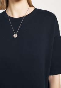 Personne portant un pull en maille bleu marine foncé et un collier pendentif rond en argent sur une fine chaîne sur un fond uni.