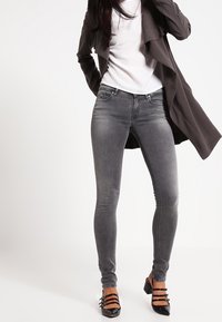 Jeans skinny grises con una textura suave, combinados con una blusa blanca y un abrigo largo gris. Tacones de charol negros con detalles a rayas.