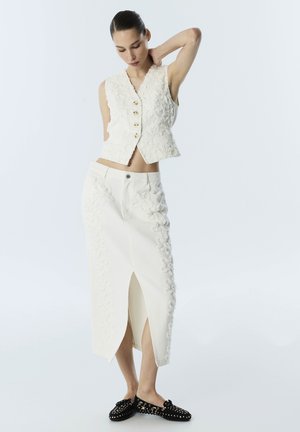 Femme portant un gilet sans manches blanc à texture florale et une jupe midi blanche assortie avec une fente à l'avant, posant avec la main derrière la nuque.