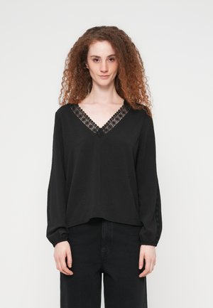 Γυναίκα με καστανά σγουρά μαλλιά που φοράει μαύρο μπλουζάκι με μακριά μανίκια και δαντέλα στο V-λαιμό και τα μανίκια, συνδυασμένο με μαύρο παντελόνι.