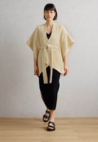 Beige poncho anysoitu, me sleeves fupi, iliyofungwa kiunoni, imepangwa juu ya mavazi meusi yenye slit upande na kuvaa na sanda za rangi ya mweusi.