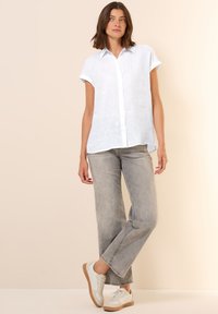 Camicia bianca in lino a maniche corte con abbottonatura frontale, abbinata a jeans larghi grigi chiari e sneakers bianche con dettagli beige.